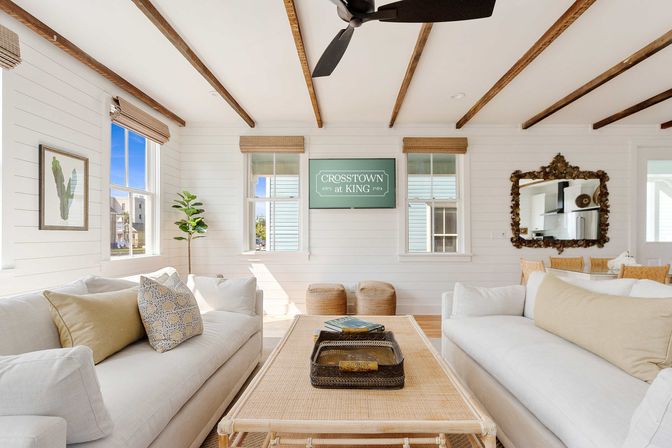 Bright coastal beach-cottage living room with white shiplap walls, exposed wooden beams, two beige sofas, rattan coffee table, woven shades and ceiling fan.
