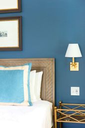Cozy bedroom corner with a woven wood headboard against a deep blue wall, turquoise velvet pillow with cream trim, white bedding, brass wall sconce with white shade, and a gold metal bedside table.