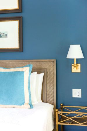 Cozy bedroom corner with a woven wood headboard against a deep blue wall, turquoise velvet pillow with cream trim, white bedding, brass wall sconce with white shade, and a gold metal bedside table.