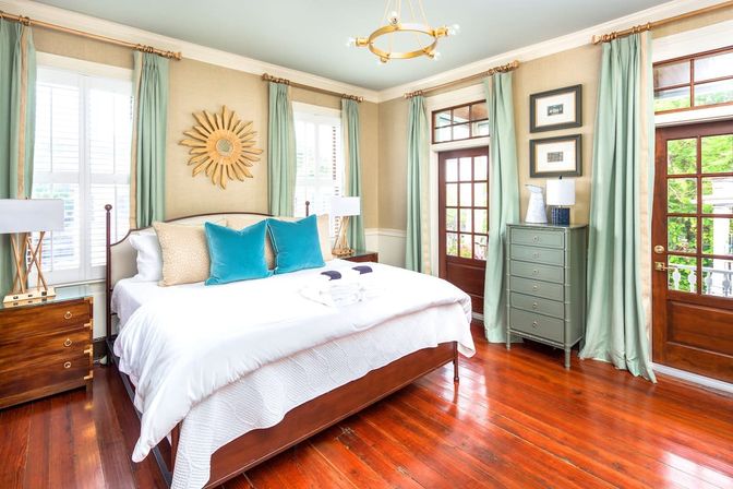 Sunlit coastal-chic bedroom with white king bed and teal accent pillows, gold sunburst mirror above the headboard, light green curtains framing windows and French doors, gray dresser and wooden nightstands on glossy hardwood floors