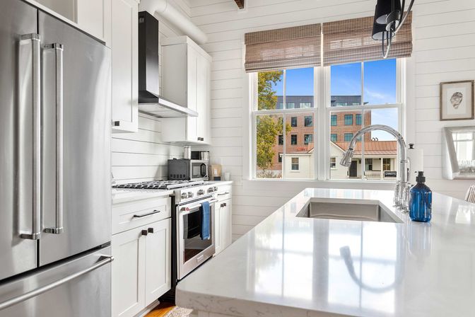 Bright modern farmhouse kitchen with white shiplap walls, quartz island and undermount sink, chrome faucet, stainless-steel fridge and range, large window with woven shades overlooking brick city buildings.