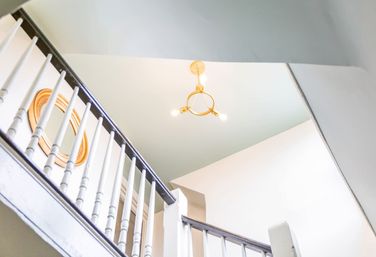 Upward view of a bright home interior with a white wooden staircase balustrade and dark handrail, pale green ceiling, round gold-framed mirror, and a brass three-bulb chandelier