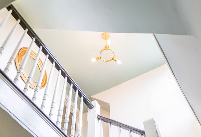 Upward view of a bright home interior with a white wooden staircase balustrade and dark handrail, pale green ceiling, round gold-framed mirror, and a brass three-bulb chandelier