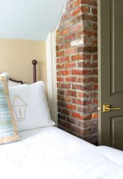 Cozy bedroom interior with white quilted bedding and patterned pillows, wooden bedpost, exposed red brick chimney next to a partially open gray door with brass handle.