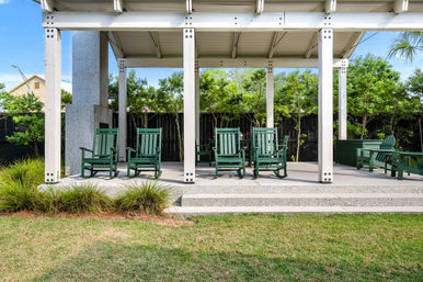 Covered backyard pavilion with white posts and concrete steps, an inviting row of green wooden rocking chairs on a concrete patio, surrounded by lawn, shrubs, and a privacy fence.