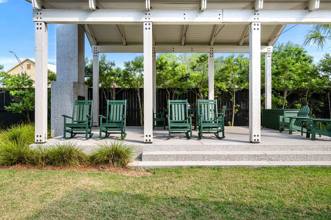 Covered backyard pavilion with white posts and concrete steps, an inviting row of green wooden rocking chairs on a concrete patio, surrounded by lawn, shrubs, and a privacy fence.