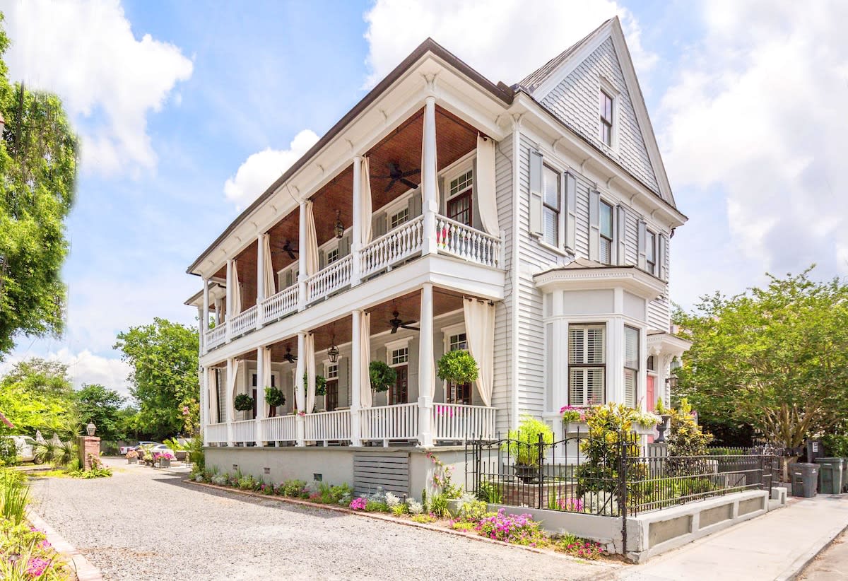 Sunlit white Victorian-style home with double wraparound porches and tall columns, bay window, hanging potted plants, iron fence and gravel driveway in a leafy neighborhood.