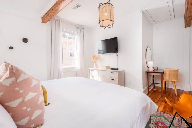 Bright modern bedroom with exposed wooden beams and hardwood floors, white bedding with a pink patterned pillow, wall-mounted TV over a dresser, woven pendant light, small wooden desk and wicker chair — light-filled, cozy minimalist design.