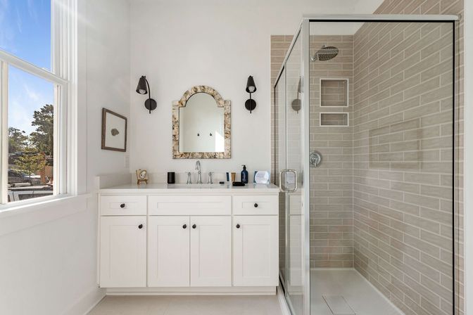 Bright sunlit bathroom with white vanity and single faucet, decorative mirror, black wall sconces, and glass-enclosed beige subway-tiled shower.