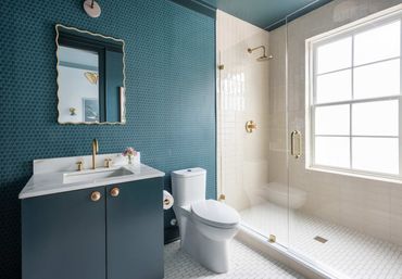 Bright modern bathroom featuring teal patterned wallpaper, marble-top vanity with gold faucet and scalloped mirror, white one-piece toilet, and glass-enclosed walk-in shower with gold fixtures and large window