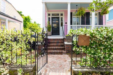 Charming pink cottage with white-columned porch, black front door, brick steps and herringbone walkway, wrought-iron gate and mailbox framed by white flowering vines and potted plants.
