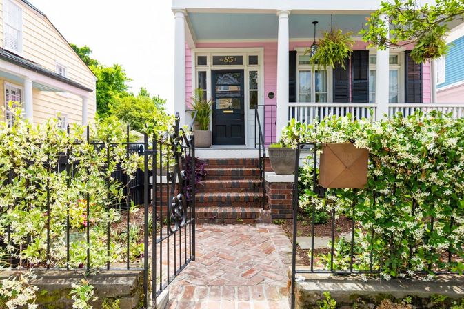 Charming pink cottage with white-columned porch, black front door, brick steps and herringbone walkway, wrought-iron gate and mailbox framed by white flowering vines and potted plants.