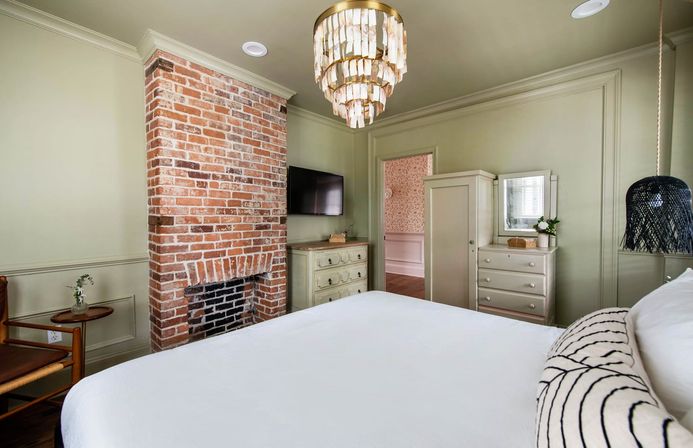 Cozy boutique-style bedroom with exposed red-brick fireplace, layered shell chandelier, sage-green walls, white bed, dresser and wall-mounted TV