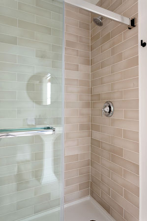 Sleek corner shower with beige subway tile, chrome rain showerhead and single-lever valve, sliding glass door with chrome handle, white shower pan and faint reflection of a pedestal sink and round mirror.