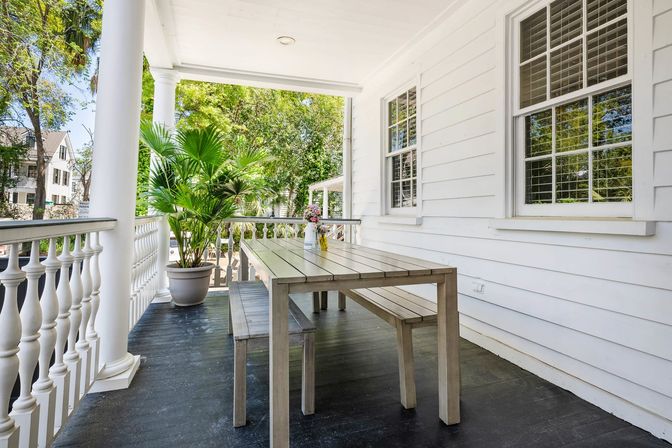 Sunlit white front porch with classical columns and balustrade, gray wooden picnic table and benches, potted palm and vase of flowers overlooking a leafy suburban street.