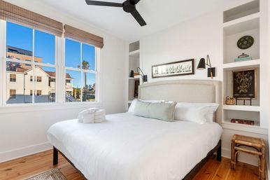 Sunlit coastal-style bedroom with a white king bed, soft green lumbar pillow, built-in shelves, hardwood floors, black ceiling fan, and a large window framing a palm tree and neighboring beach-style houses.
