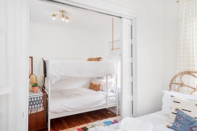Bright, light-filled small bedroom with white bunk beds, crisp white bedding, hardwood floors, colorful rug and boho accents like a rattan chair, patterned pillows and a potted plant.