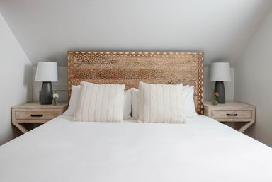 Cozy attic bedroom with sloped ceiling, carved wooden headboard, crisp white bedding, two textured cream pillows and matching light-wood bedside tables with gray lamps and small flower vases.
