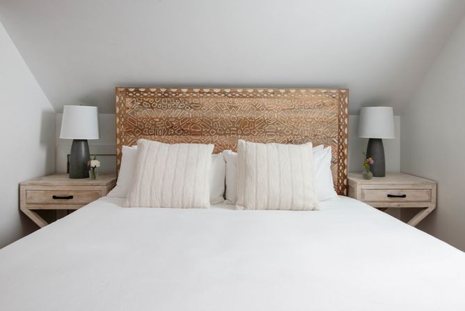 Cozy attic bedroom with sloped ceiling, carved wooden headboard, crisp white bedding, two textured cream pillows and matching light-wood bedside tables with gray lamps and small flower vases.