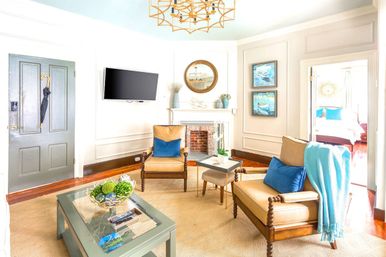 Bright sunlit living room with tan leather armchairs, blue pillows and throw, glass-top coffee table with greenery, wall-mounted TV, and small decorative fireplace in coastal-inspired decor.