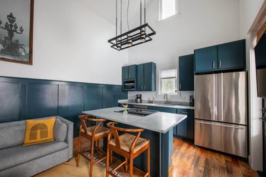 Bright modern kitchen with navy blue cabinets, marble island and breakfast bar, stainless-steel French-door refrigerator, wooden bar stools, hardwood floors, industrial pendant light and gray sofa with yellow accent pillow.