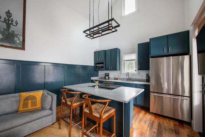 Bright modern kitchen with navy blue cabinets, marble island and breakfast bar, stainless-steel French-door refrigerator, wooden bar stools, hardwood floors, industrial pendant light and gray sofa with yellow accent pillow.