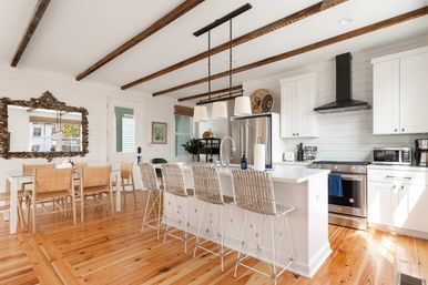 Bright coastal farmhouse kitchen with exposed wood ceiling beams, white shiplap walls, large island with pendant lights and wicker bar stools, stainless steel appliances, dining table, and warm pine floors.