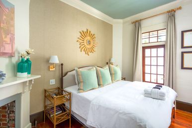 Bright coastal-chic bedroom with white bedding, mint green pillows, gold sunburst mirror above the headboard, teal vases on the mantel, French doors and hardwood floors.
