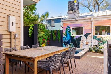 Sunny urban backyard patio with wooden dining table and six modern woven chairs on brick pavers, vertical green wall, small plunge pool and vibrant peacock-and-flower mural on the exterior wall.