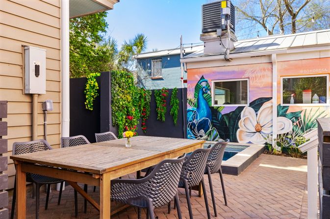 Sunny urban backyard patio with wooden dining table and six modern woven chairs on brick pavers, vertical green wall, small plunge pool and vibrant peacock-and-flower mural on the exterior wall.