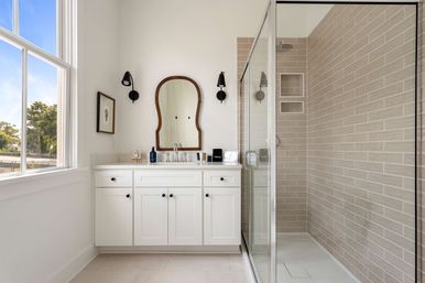 Bright modern bathroom with white shaker vanity, arched wood-framed mirror, black wall sconces, glass-enclosed beige subway-tile shower, and large window flooding the space with natural light.
