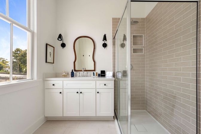 Bright modern bathroom with white shaker vanity, arched wood-framed mirror, black wall sconces, glass-enclosed beige subway-tile shower, and large window flooding the space with natural light.