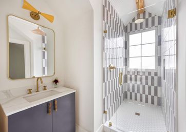 Sunlit modern bathroom with navy marble vanity, brass fixtures and rounded mirror, yellow sconce, plus a glass shower featuring vertical gray-and-white striped tiles and white hexagon floor tiles by a window.