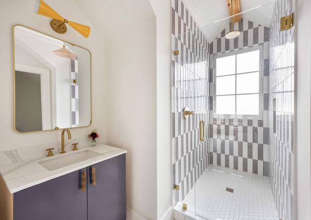 Sunlit modern bathroom with navy marble vanity, brass fixtures and rounded mirror, yellow sconce, plus a glass shower featuring vertical gray-and-white striped tiles and white hexagon floor tiles by a window.