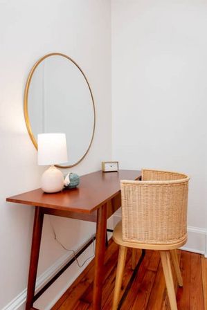Cozy home office nook with a mid-century wooden desk, woven rattan chair, round gold-framed mirror, white table lamp and warm hardwood floors.