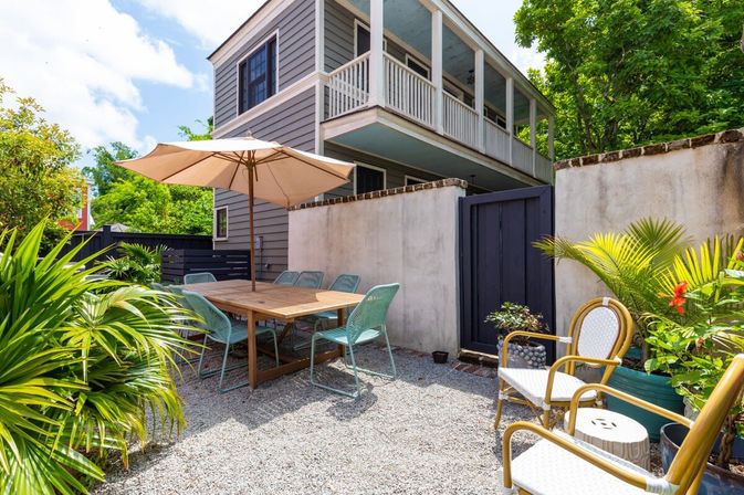 Sunny backyard patio with umbrella-shaded wooden dining table and teal chairs on a gravel courtyard, tropical potted palms, and a two-story gray house with a covered balcony — inviting outdoor dining.