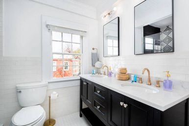 Bright modern bathroom with black double vanity and marble countertop, gold faucets, dual mirrors, white subway tile and a window showing a red building outside.