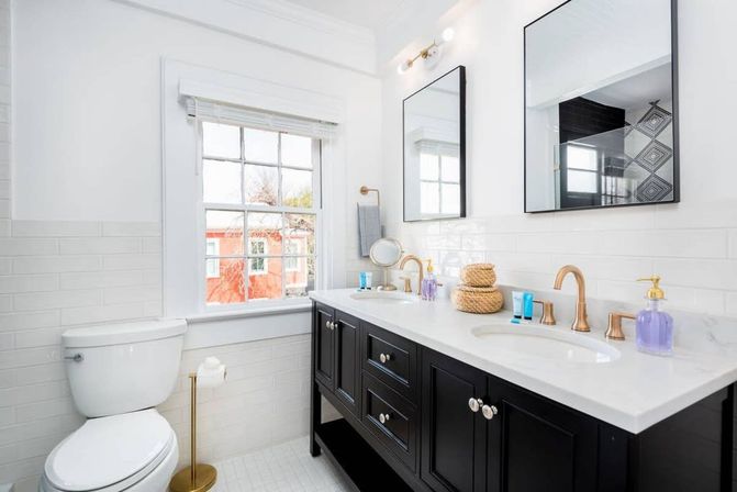 Bright modern bathroom with black double vanity and marble countertop, gold faucets, dual mirrors, white subway tile and a window showing a red building outside.