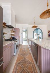 Bright modern galley kitchen with mauve cabinets, terrazzo countertops, gold faucet and rattan pendant lights; patterned runner rug leads to a blue arched doorway.