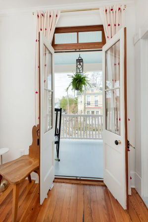 Bright entry with open French doors leading to a covered front porch with white railing, hanging fern lantern, hardwood floors, and colorful Victorian houses across the street.
