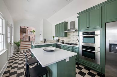 Bright modern kitchen with sage-green cabinets, white quartz island with bar stools, black-and-white checkerboard floor, stainless steel double ovens and refrigerator, patterned tile backsplash and large windows