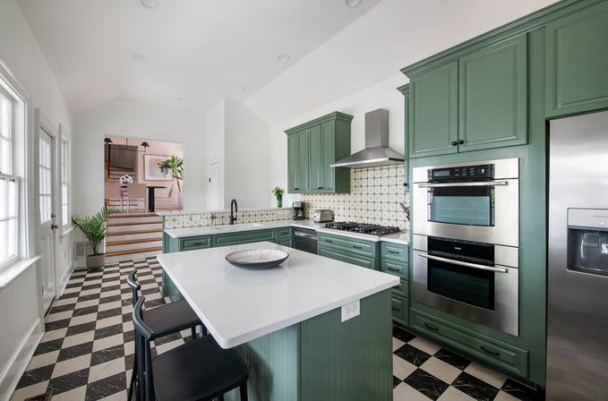 Bright modern kitchen with sage-green cabinets, white quartz island with bar stools, black-and-white checkerboard floor, stainless steel double ovens and refrigerator, patterned tile backsplash and large windows