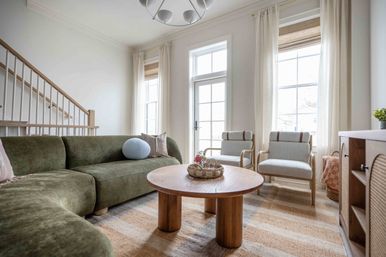 Bright sunlit living room with an olive-green sectional, round wooden coffee table with woven tray, two light upholstered armchairs, tall grid windows with sheer curtains, neutral striped rug and wooden staircase — cozy modern interior