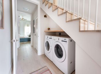 Bright under-stair laundry nook with white front-load washer and dryer, light wood floating shelf and staircase, airy hallway leading to a bedroom in a modern neutral home