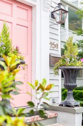 Cheerful pink front door on a white clapboard home entrance with brass house number 154, antique-style lantern, and tall urn planters overflowing with greenery and purple flowers.