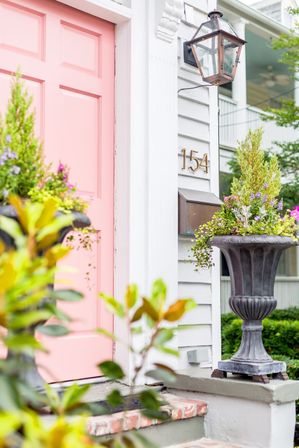 Cheerful pink front door on a white clapboard home entrance with brass house number 154, antique-style lantern, and tall urn planters overflowing with greenery and purple flowers.