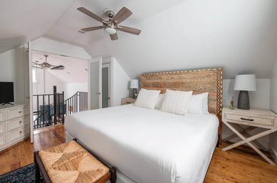 Bright loft-style master bedroom with king bed, carved wooden headboard, crisp white linens, matching nightstands and lamps, woven bench, ceiling fan and hardwood floors overlooking an open stair railing.