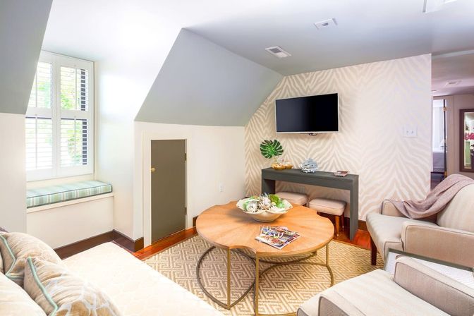 Bright cozy attic living room with sloped ceiling and window seat, round wooden coffee table, wall-mounted TV, neutral modern decor and small storage door