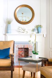 Bright coastal-style living room interior with a round brass mirror over a white mantel and exposed brick fireplace, tan leather armchairs with a blue pillow, a side table with a potted white orchid, and blue decorative vases.