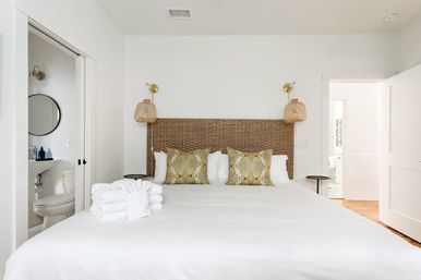 Bright, minimalist coastal-inspired bedroom with white linens, woven rattan headboard, green patterned throw pillows, hanging wicker sconces, en-suite bathroom visible and neatly folded white towels on the bed.